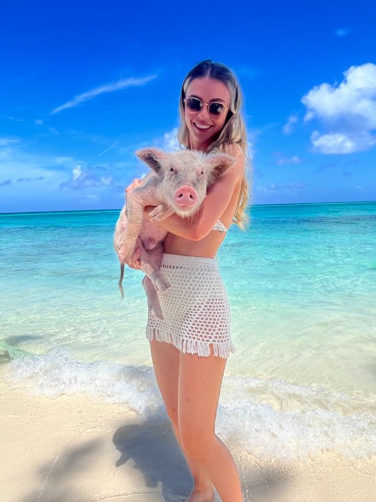 A stunning girl stands on a sandy beach in the Bahamas, holding a pig in her hand